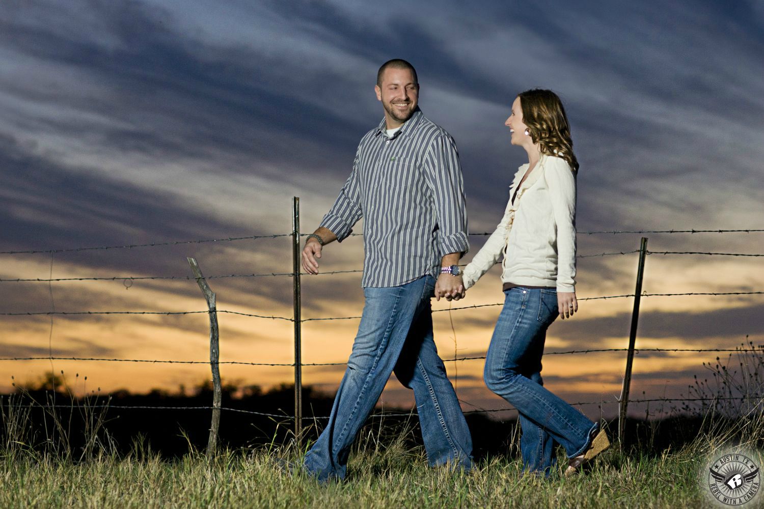 Guy with short cut dark hair wearing a striped white and grey button up dress shirt and blue jeans leads a dirty blond haired girl wearing a white low cut button down sweater with blue jeans along a fence in front of a cloudy orange and blue and grey sunset near Georgetown Texas in this colorful engagement photograph in rural Austin, Texas.